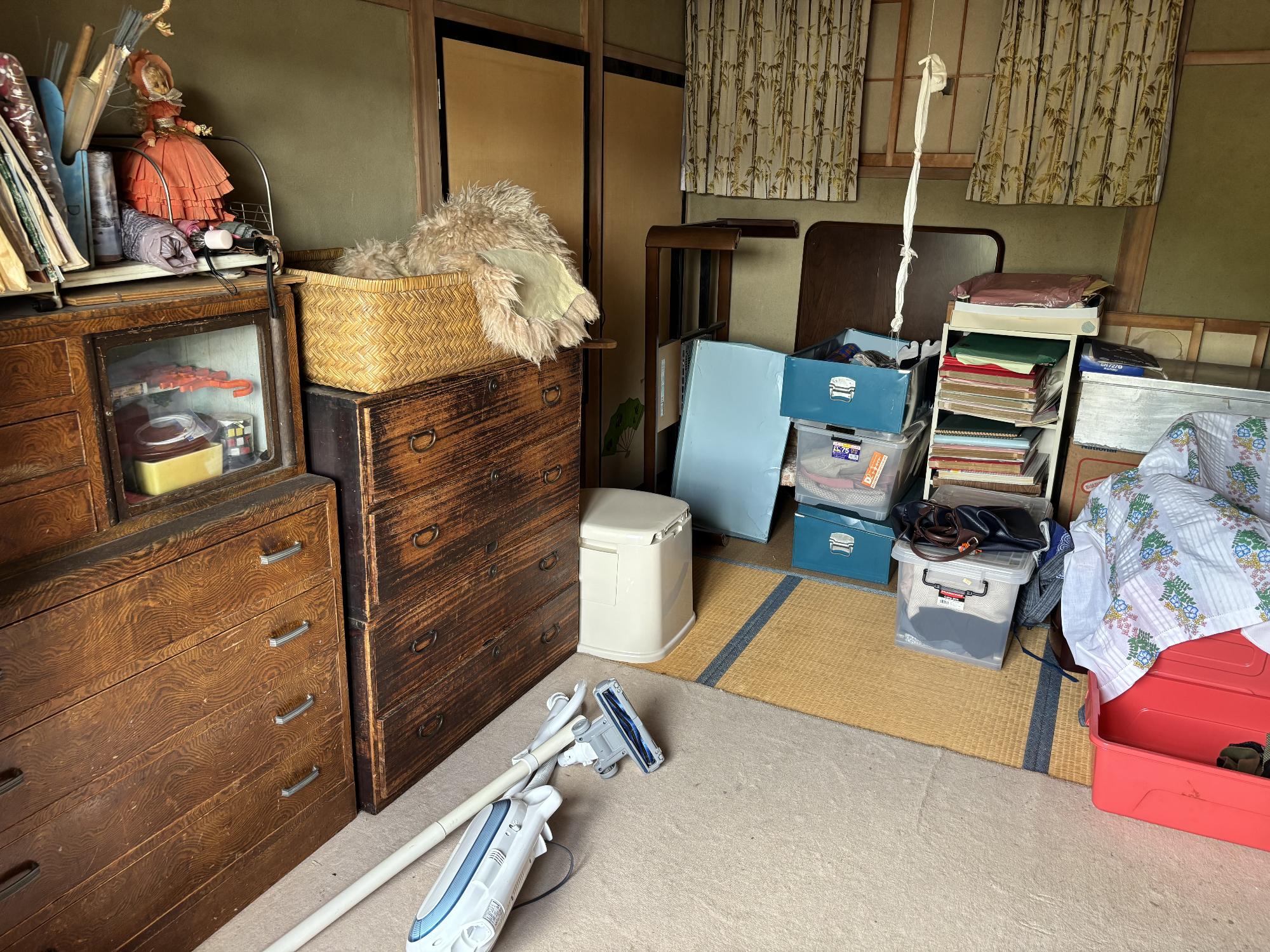 左側にタンス、正面奥上部にカーテンが半開きの障子戸、左側奥に押し入れ、畳の上にカーペットを引き、その上に掃除機、座卓、収納ケースを置いてある8帖和室内見の写真