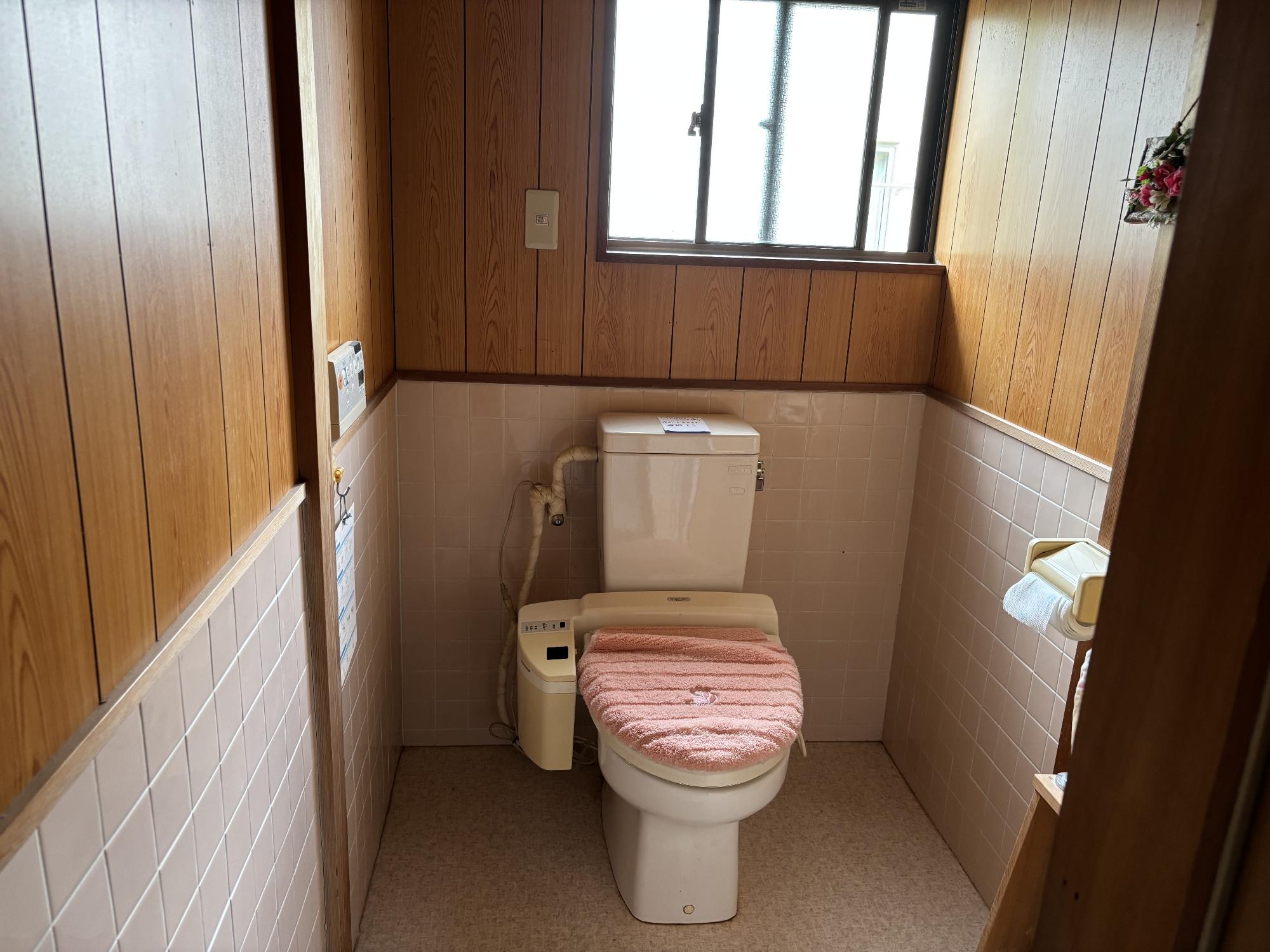 右側にトイレットペーパーホルダー、左側にリモコン、正面中央にタンク付き様式便座、正面中央上部に曇りガラスのサッシがあるトイレの写真