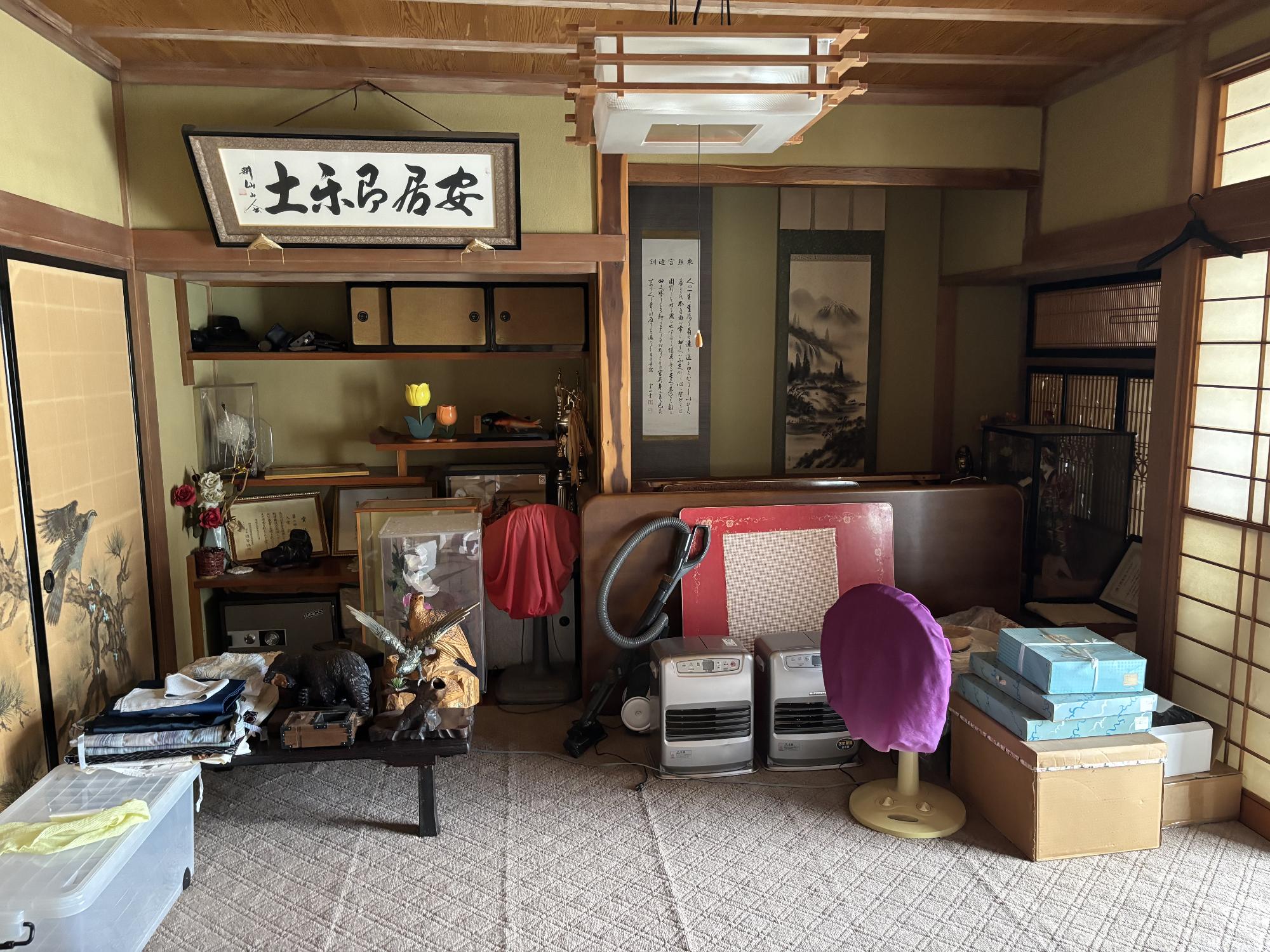 左側に襖引き戸、右側に障子戸、正面奥右側に書院作りの床の間、正面奥左側に床の間、畳の上にカーペットを引き、その上にテーブル、ファンヒーター、扇風機を置いてある8帖和室内観の写真