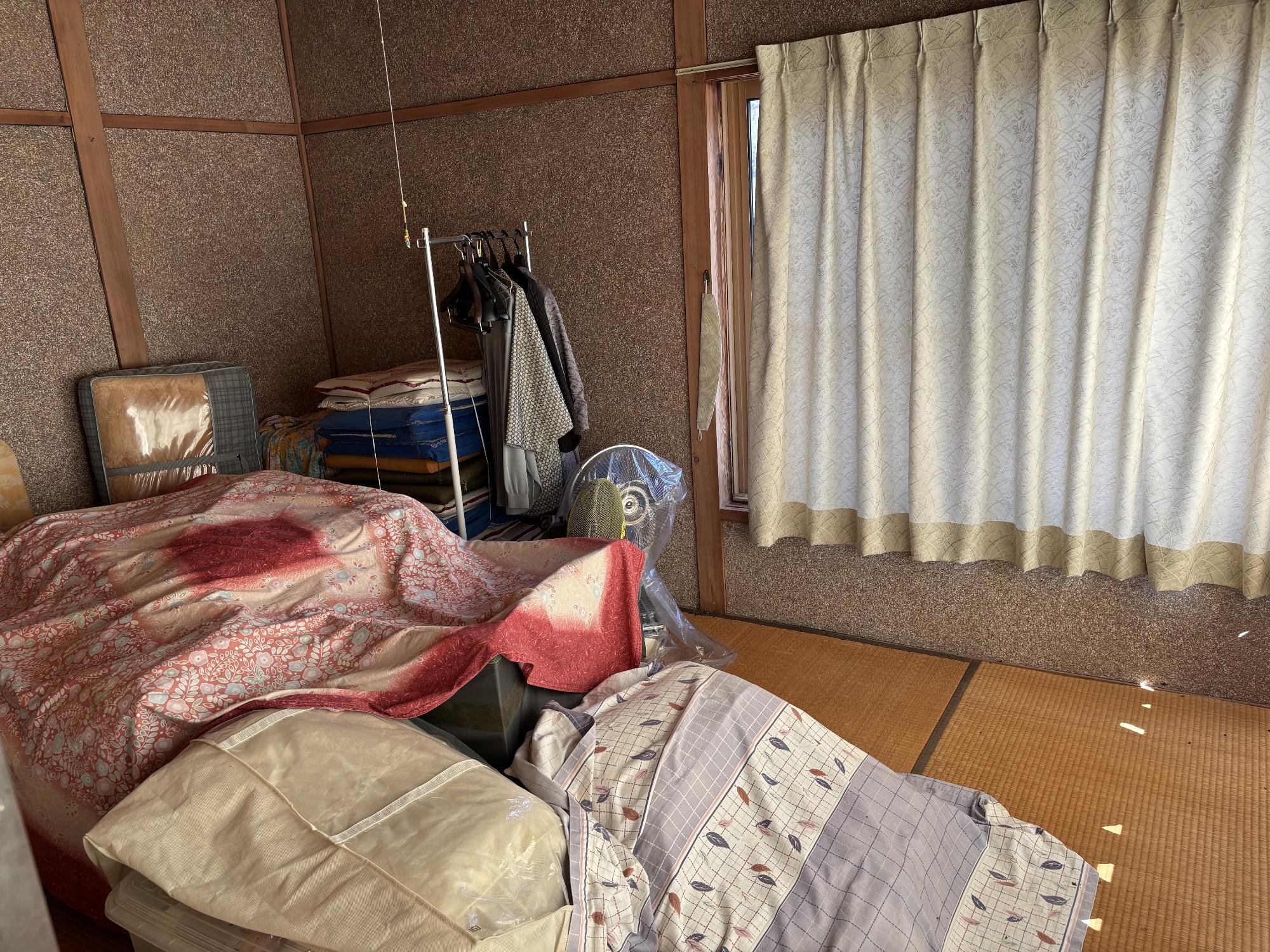右側奥にカーテンを閉じたアルミサッシ戸、正面奥に扇風機、移動式ハンガー、正面手前に布団類を重ねてある、6帖和室内見の写真