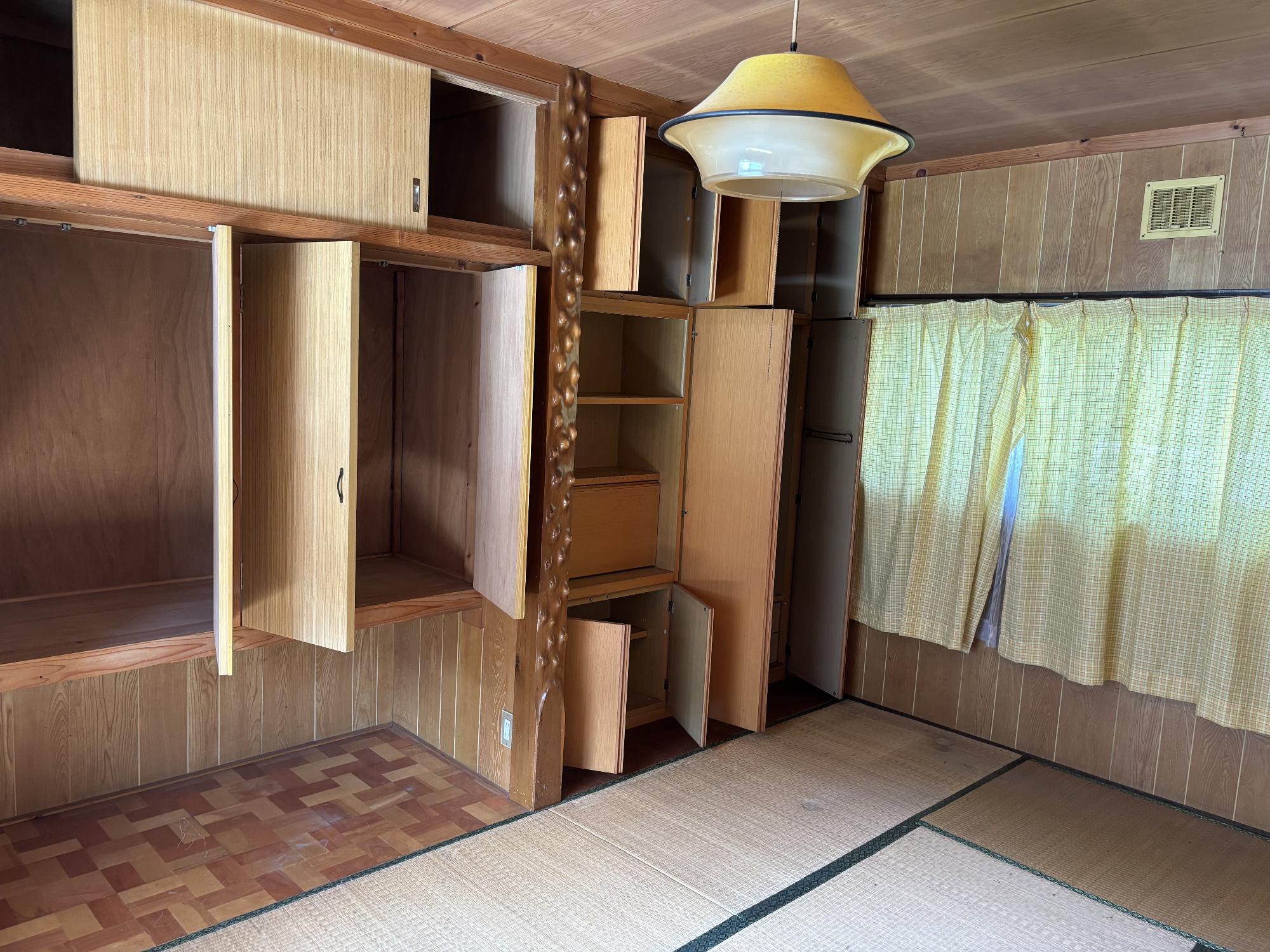 正面奥中央に床柱、その左右に収納家具、右側奥に閉じたカーテン、中央上部に照明器具ある7.5帖和室内見の写真