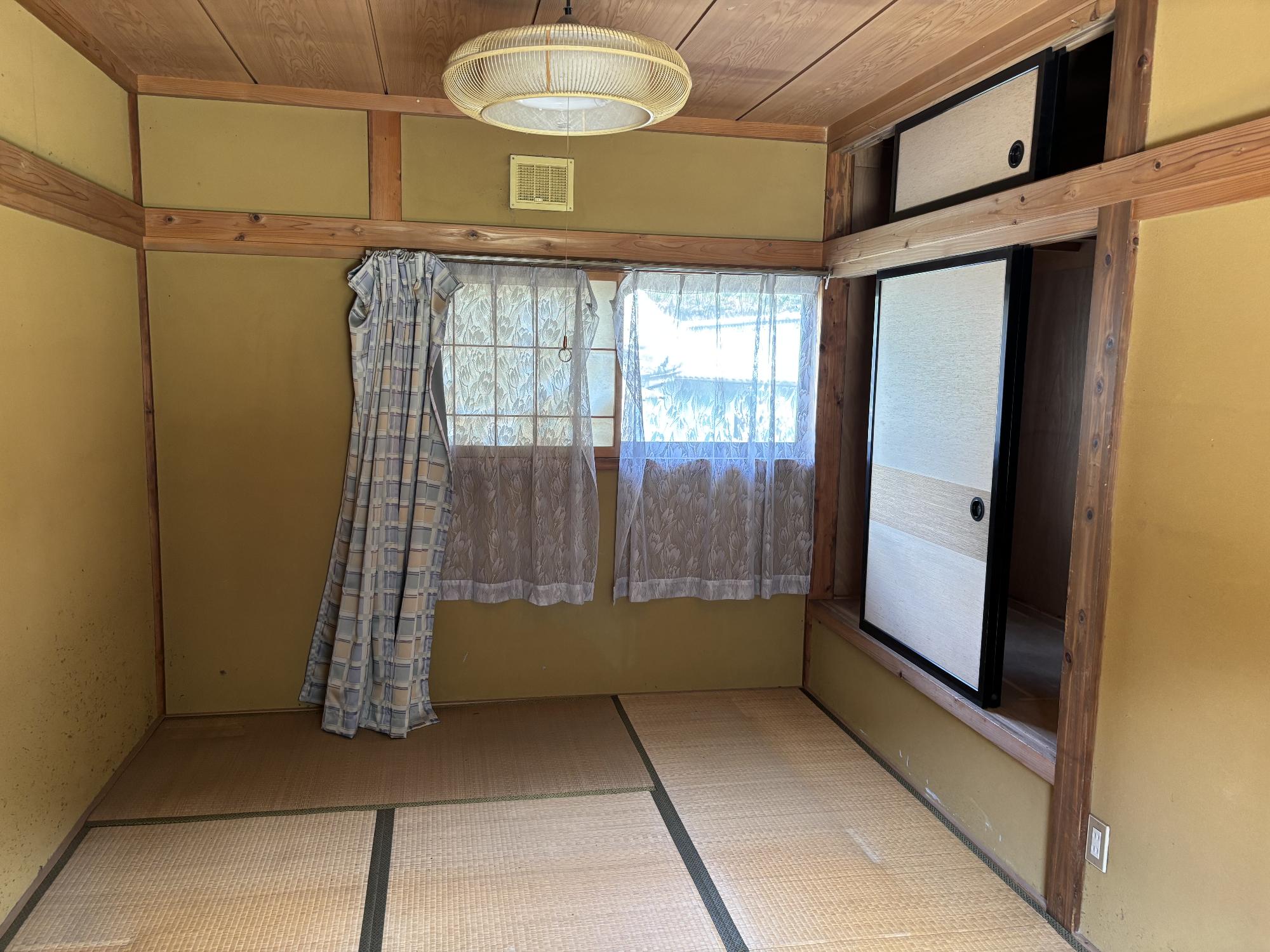 正面奥左側に閉じたカーテン、正面奥中央に閉じたレースのカーテン、右側に押し入れ、正面中央上部に照明器具ある6帖和室内観の写真