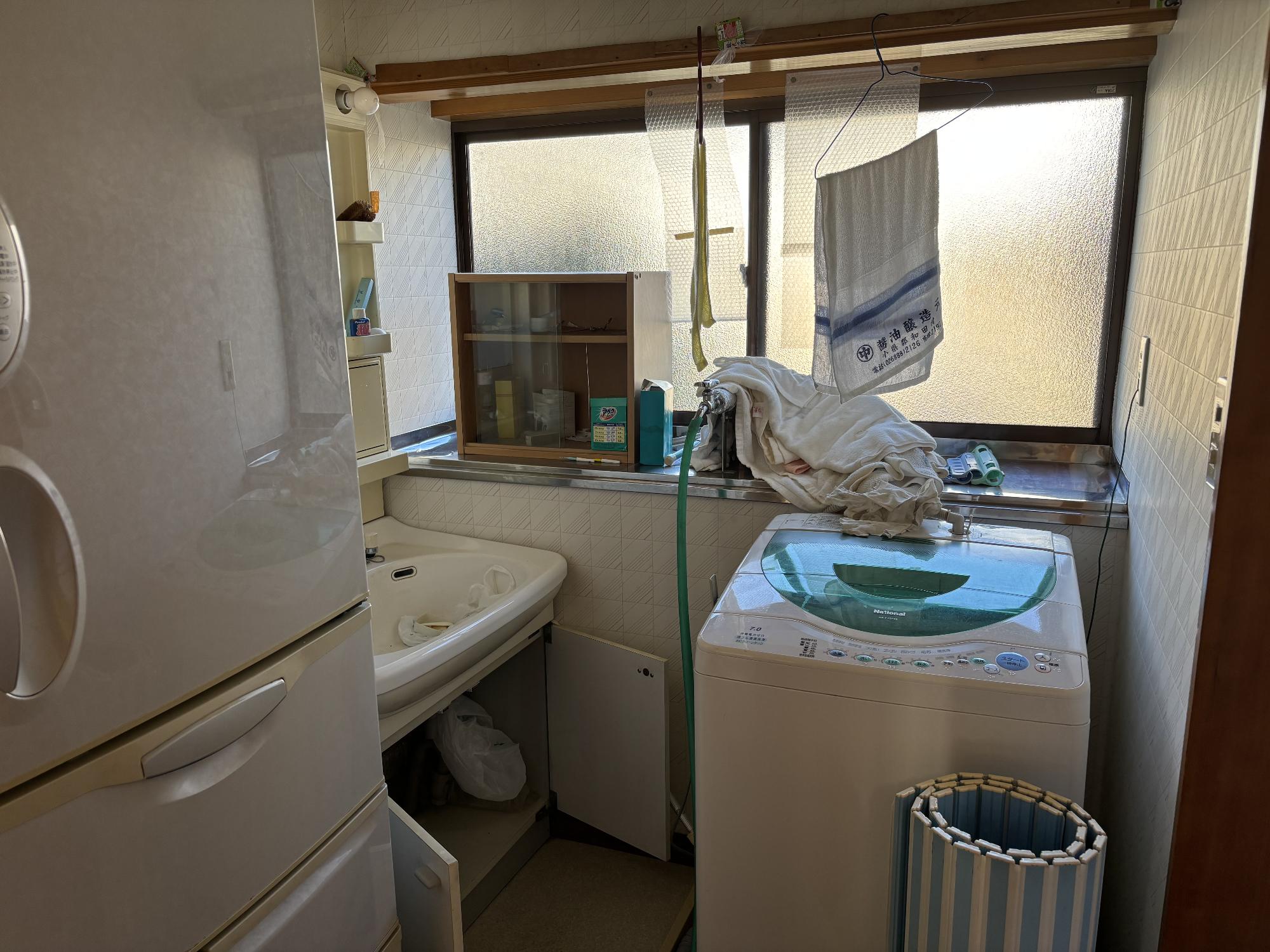 左側に冷蔵庫、その奥に収納ミラー付洗面台、正面奥上部にアルミ曇りガラスサッシ、正面奥右側に洗濯機、その手前に丸めた浴槽の蓋が置いてある洗面所の写真