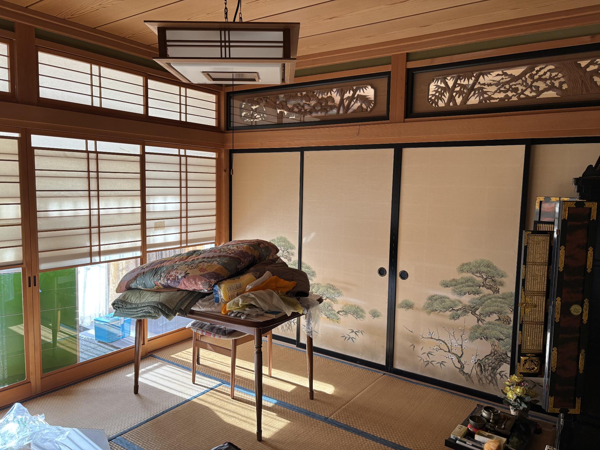 左側に仏壇、右側に雪見障子戸、正面奥に襖、欄間、中央に布団を乗せたテーブル、上部に照明器具がある8帖和室の内観写真