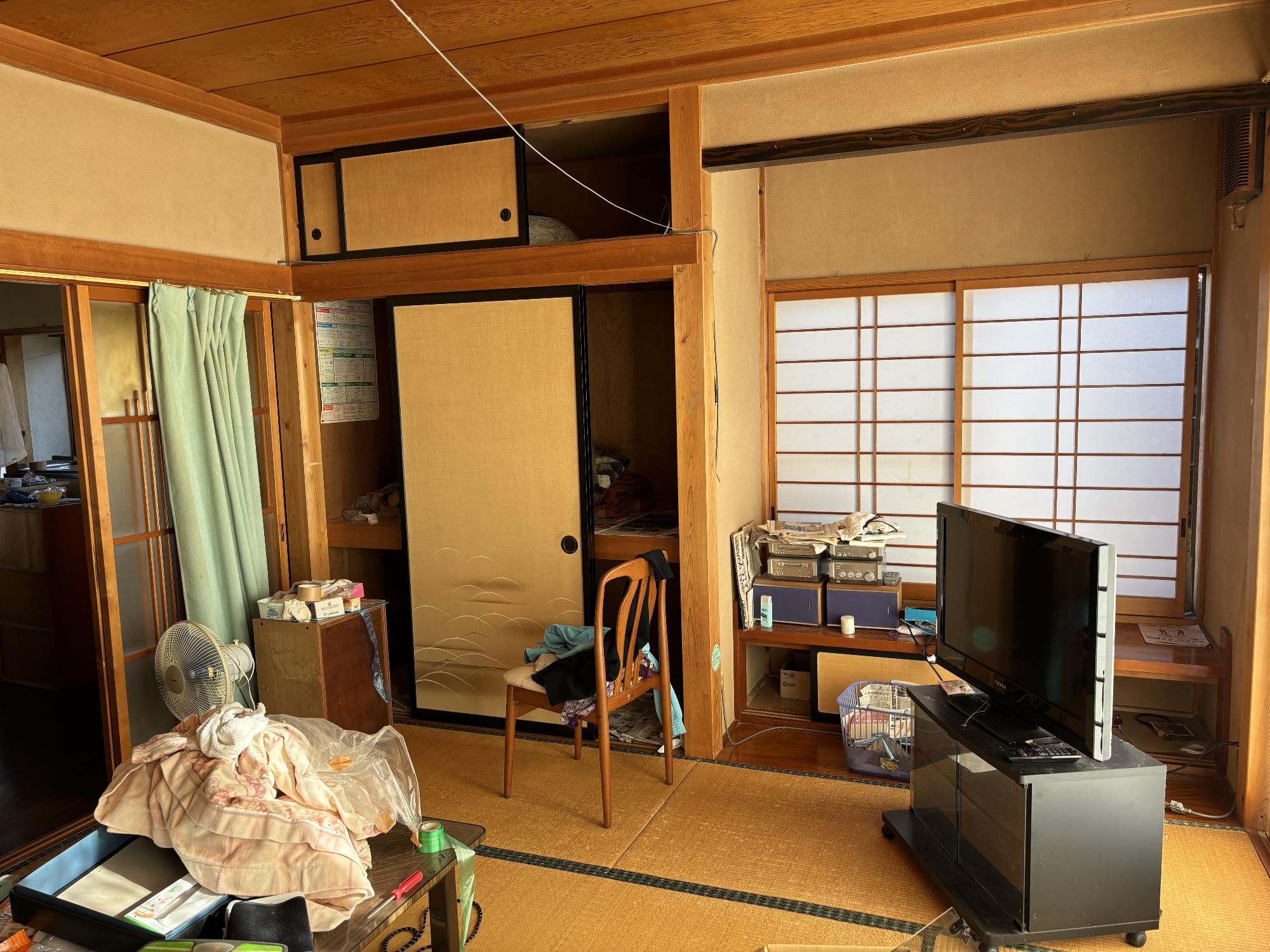 正面奥に書院作りの床の間、右側にテレビ台に乗ったテレビ、左側にガラス引き戸、布団が乗せてあるテーブル、椅子、扇風機がある、8帖和室内観の写真