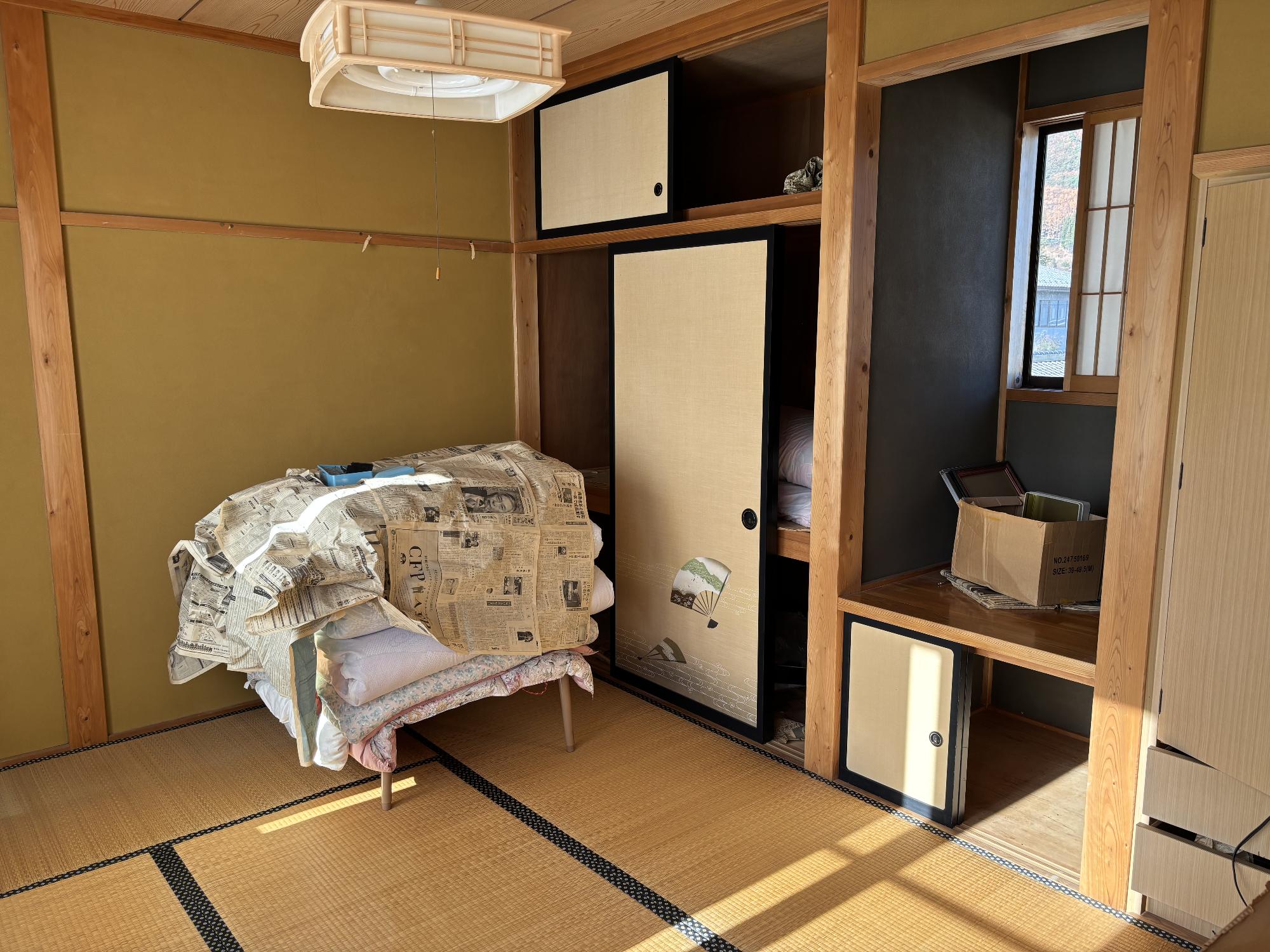 正面奥に押し入れ、その右側に床の間、クローゼット、左側に新聞紙を被せた布団が乗っているテーブル、上部に照明器具がある6帖和室内観の写真