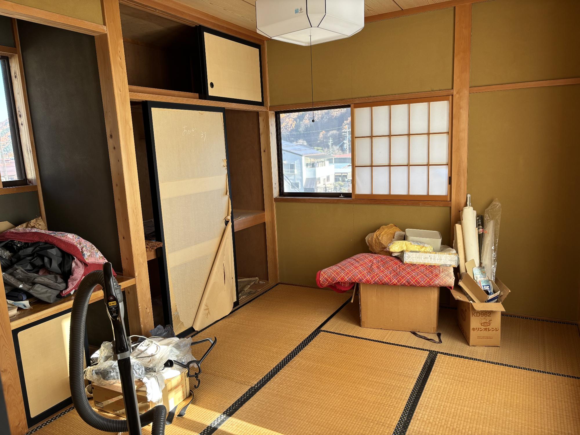 正面奥左側に押し入れ、その左側に床の間、正面奥に開いた障子戸、その手前に荷物、左側手前に掃除機が置いてある6帖和室内見の写真