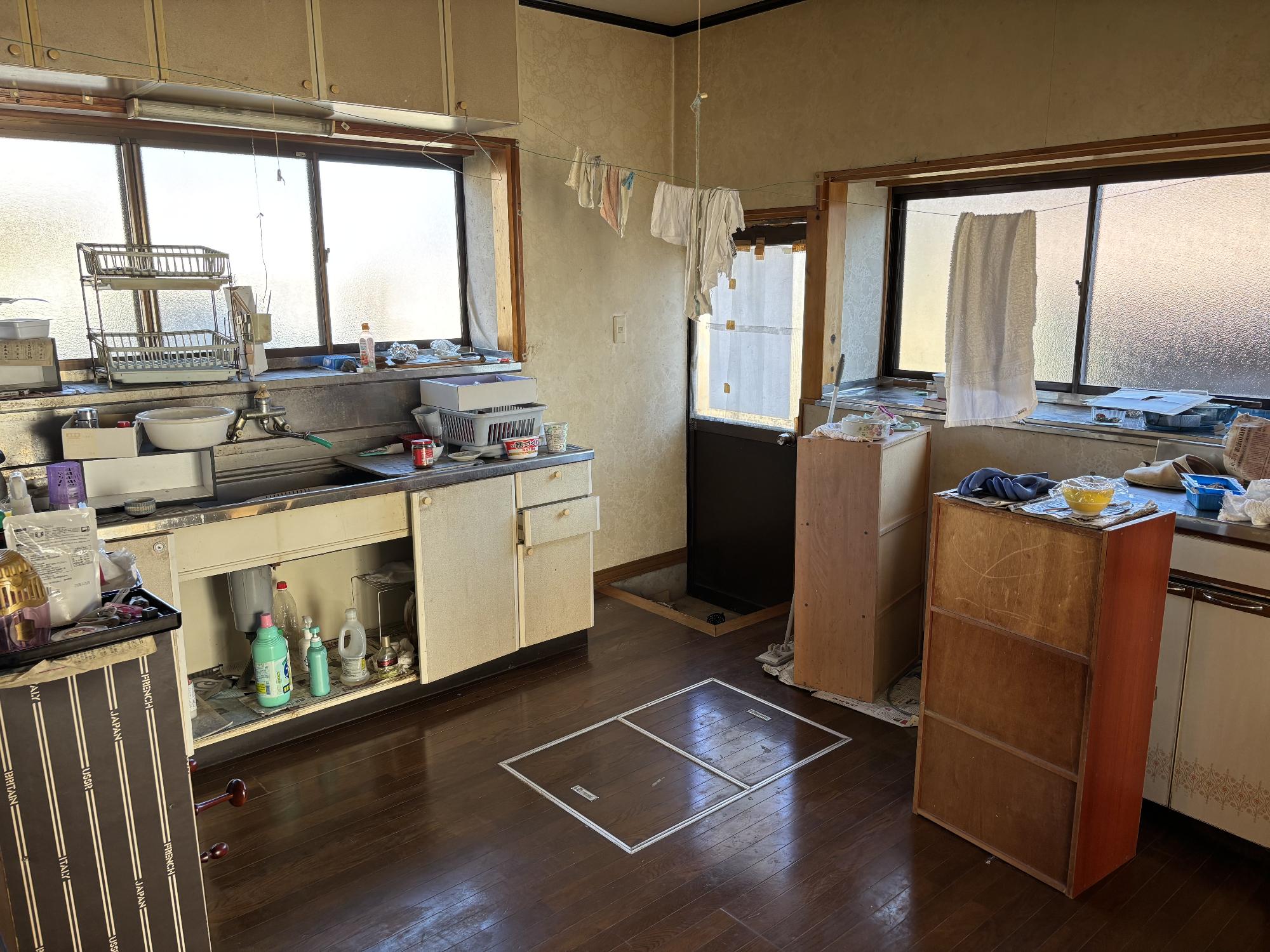 左側にシステムキッチン、その上にアルミ曇りガラスサッシ、正面奥に裏口扉、右側にカラーボックス、収納スペース、その上にアルミ曇りガラスサッシ、正面手前に床下収納庫、正面上部に手ぬぐいが干してある台所の写真