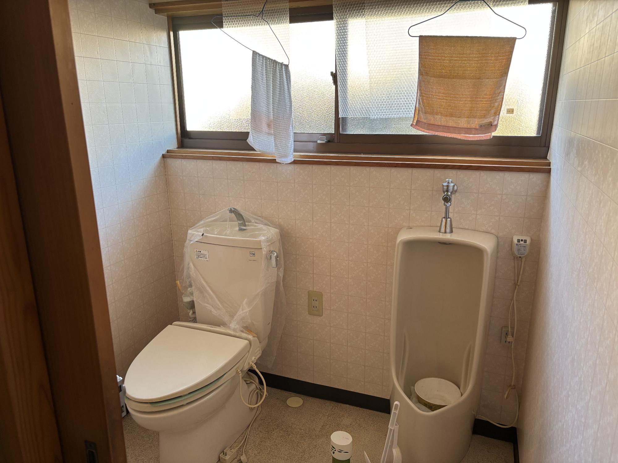 左側に洋式便座、右側に男子用便器、正面奥上部にアルミ曇りガラスサッシ、左右上部から手拭き用タオルが掛かっているトイレの写真