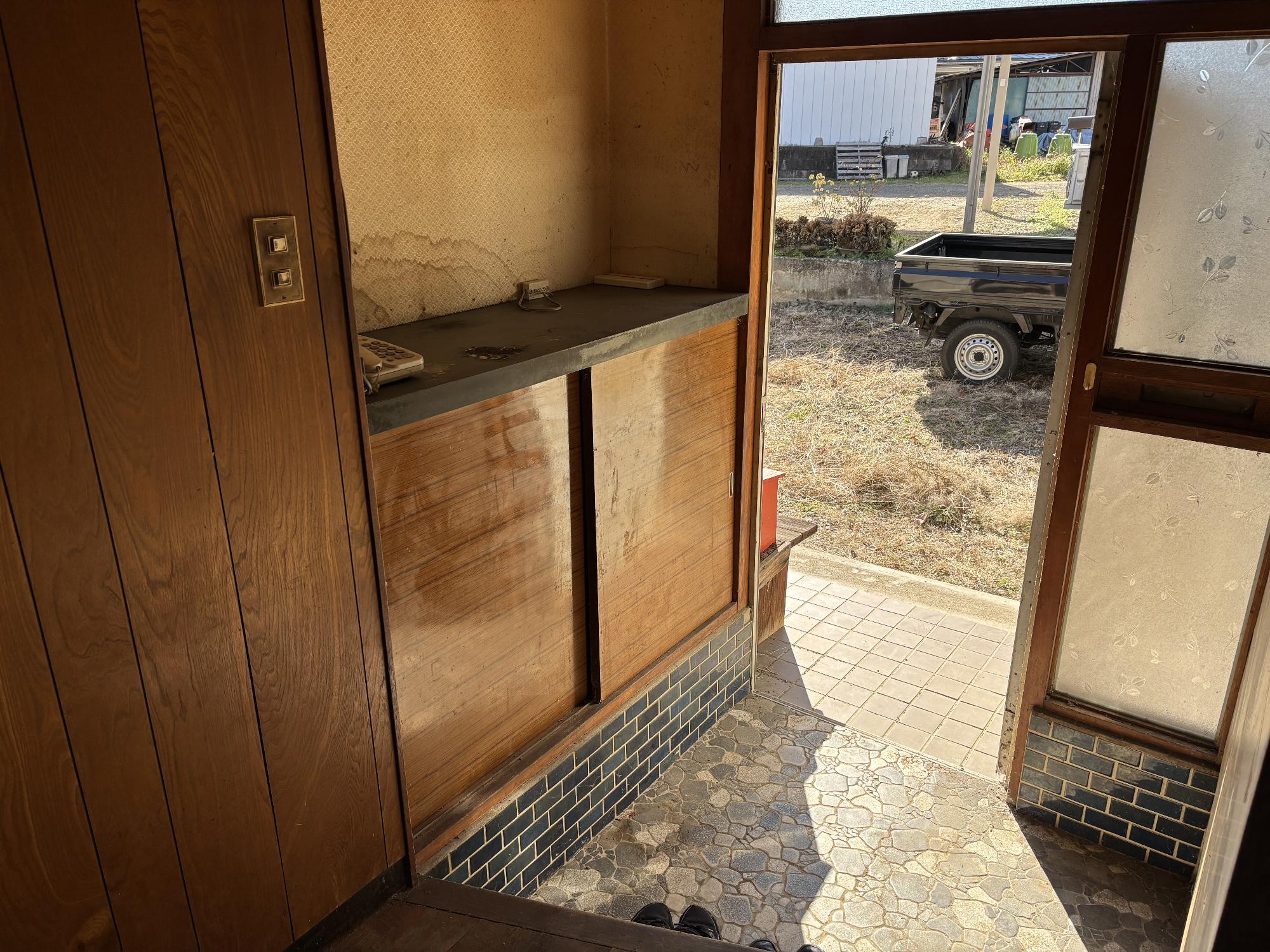 左側に下駄箱、正面中央に開いた玄関アルミサッシがある玄関内観写真