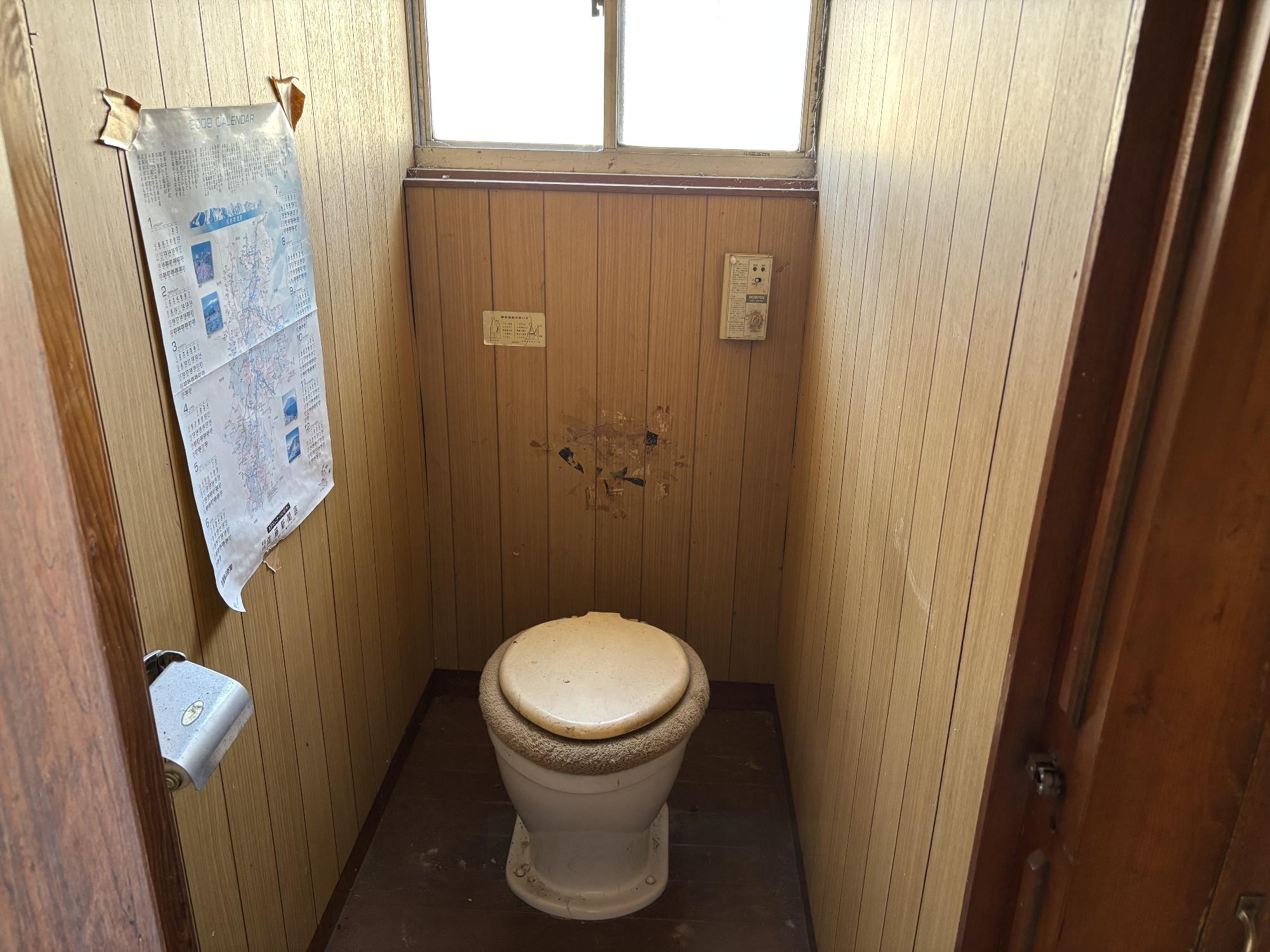 正面中央奥上部にアルミ曇りガラスサッシ、中央正面手前に洋式便座、左側にポスター、トイレットペーパーホルダーがあるトイレの写真