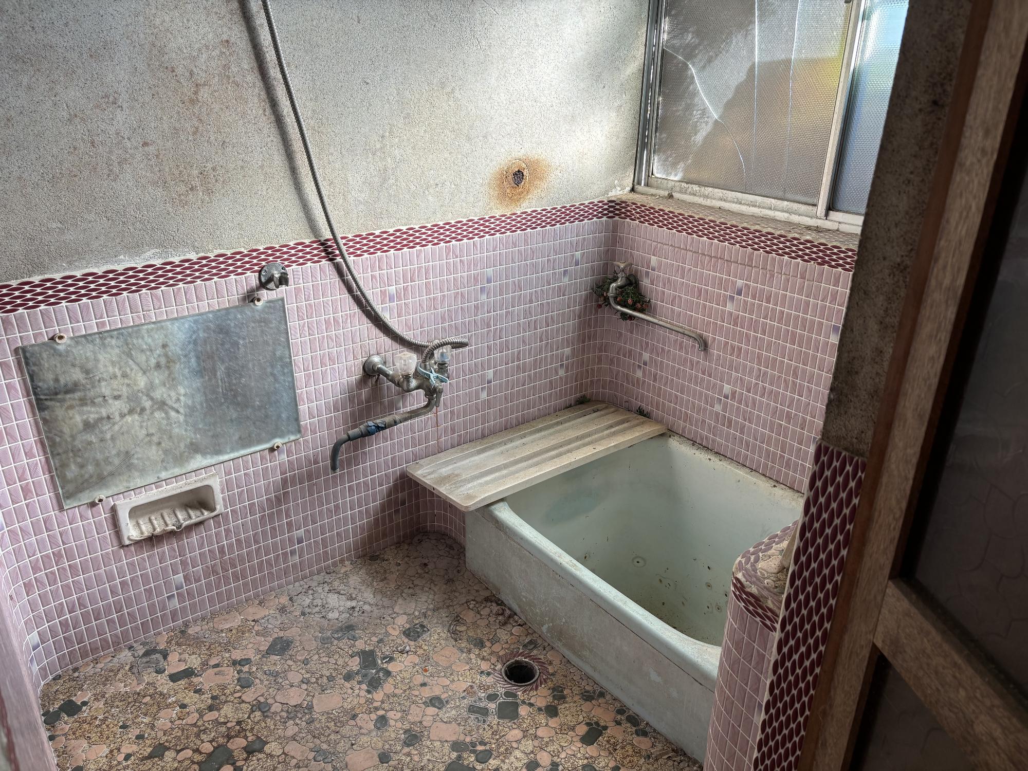 右側奥上部に曇りガラスアルミサッシ、右側に浴槽、左側に鏡、中央奥に蛇口がある、グレーのタイル張り浴室の写真