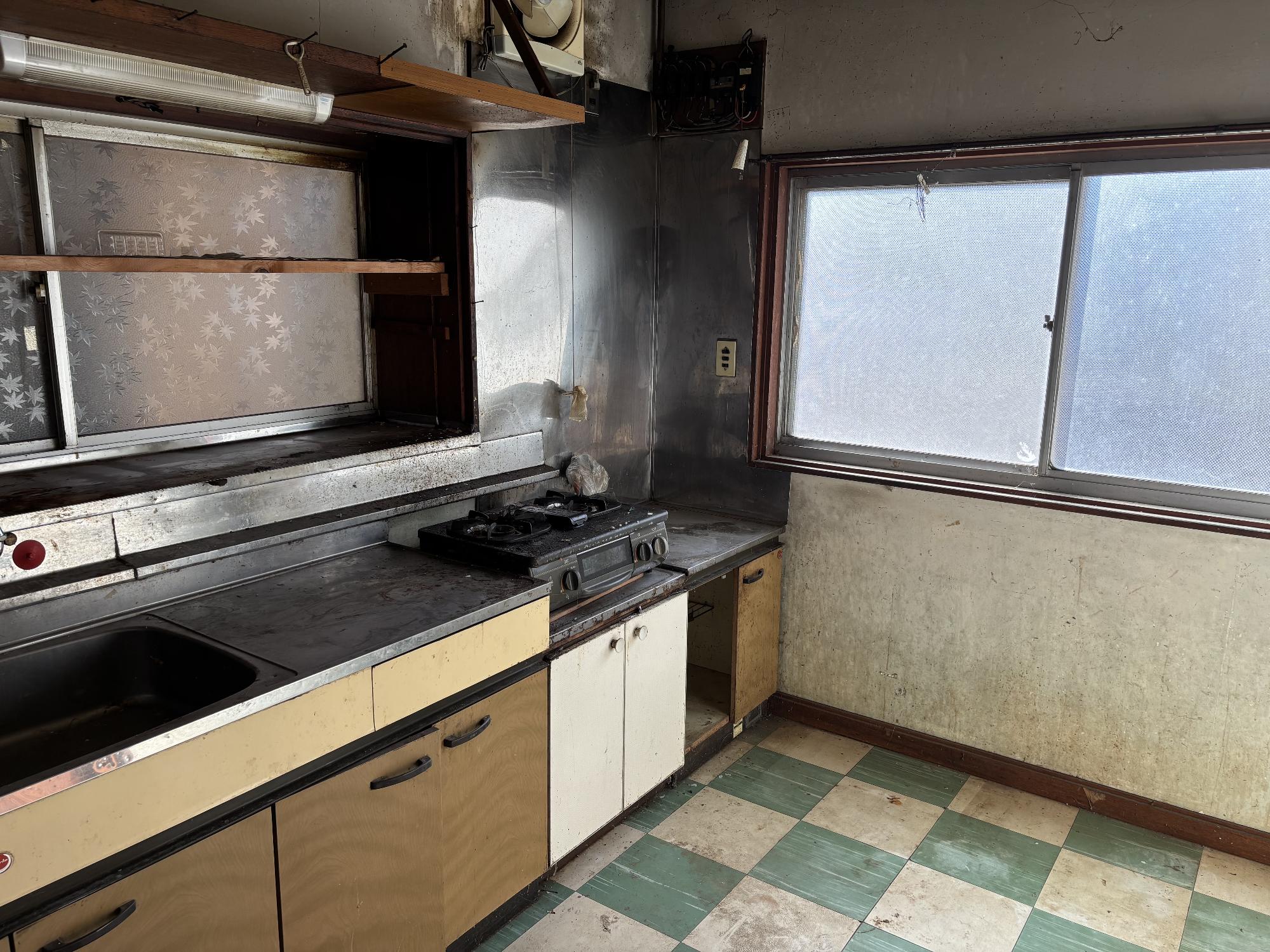 右側奥中央に曇りガラスアルミサッシ戸、左側にシンク、ガスコンロ、その上に曇りガラスサッシ戸、照明器具、換気扇がある台所の写真