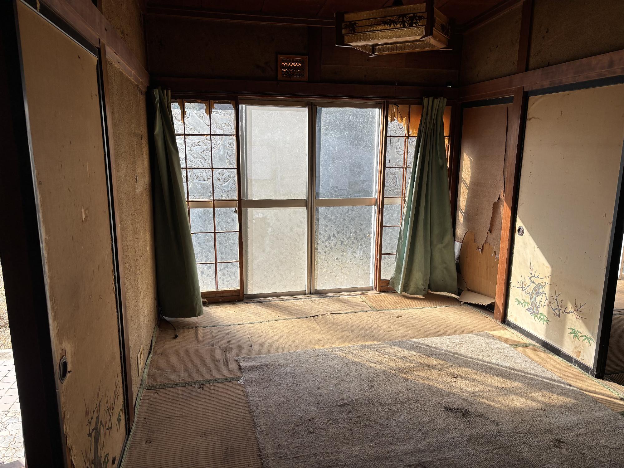 正面奥にアルミサッシ戸、その左右に開いたカーテン、左側に壁、右側に襖、正面中央上部に照明器具がある1階6帖和室の内観写真