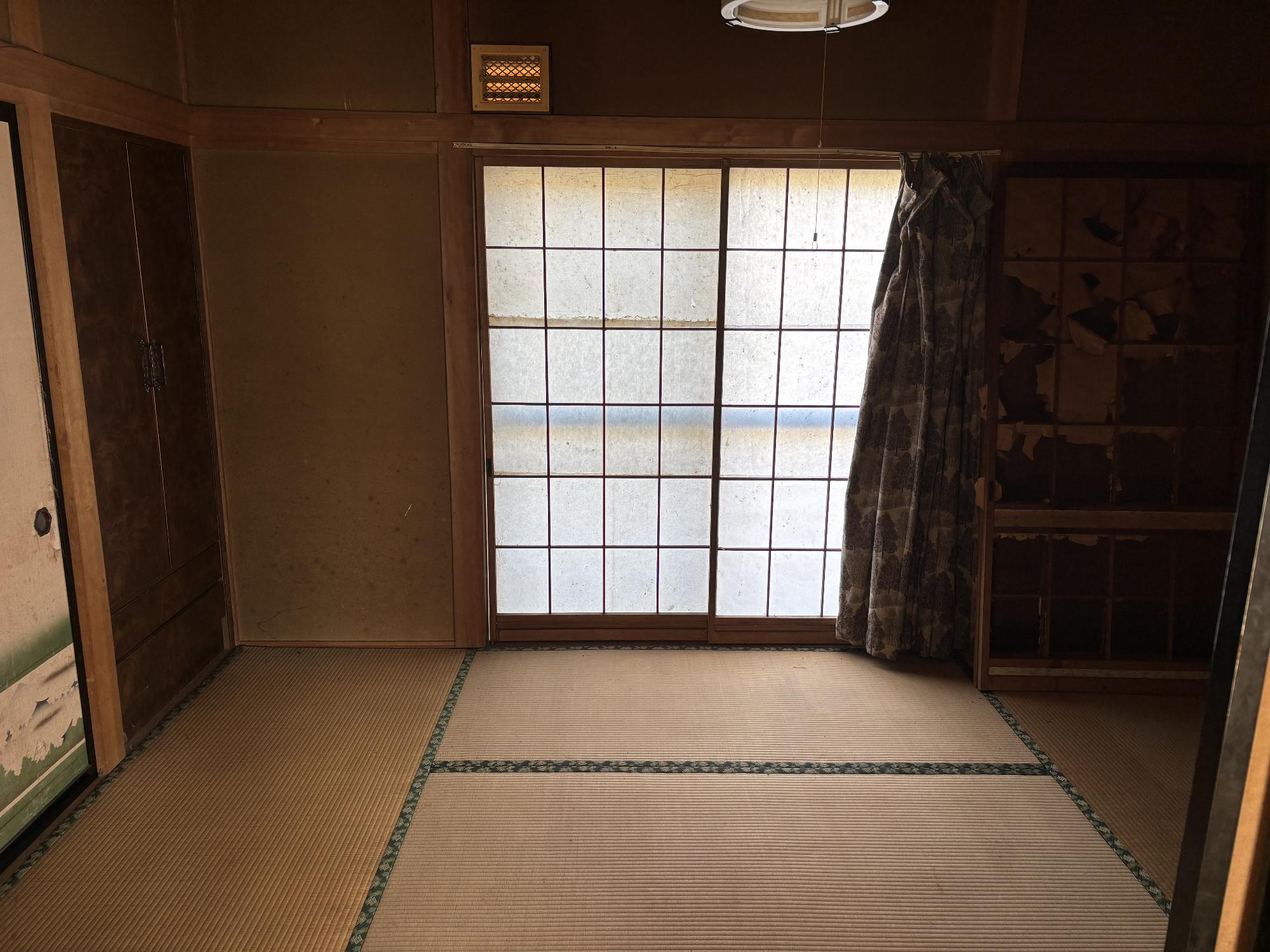 正面奥に障子戸、その右側に閉じたカーテン、左側に洋服入れ、押入れ、右側奥に机、正面中央上部に照明器具がある、1階6帖和室内観の写真