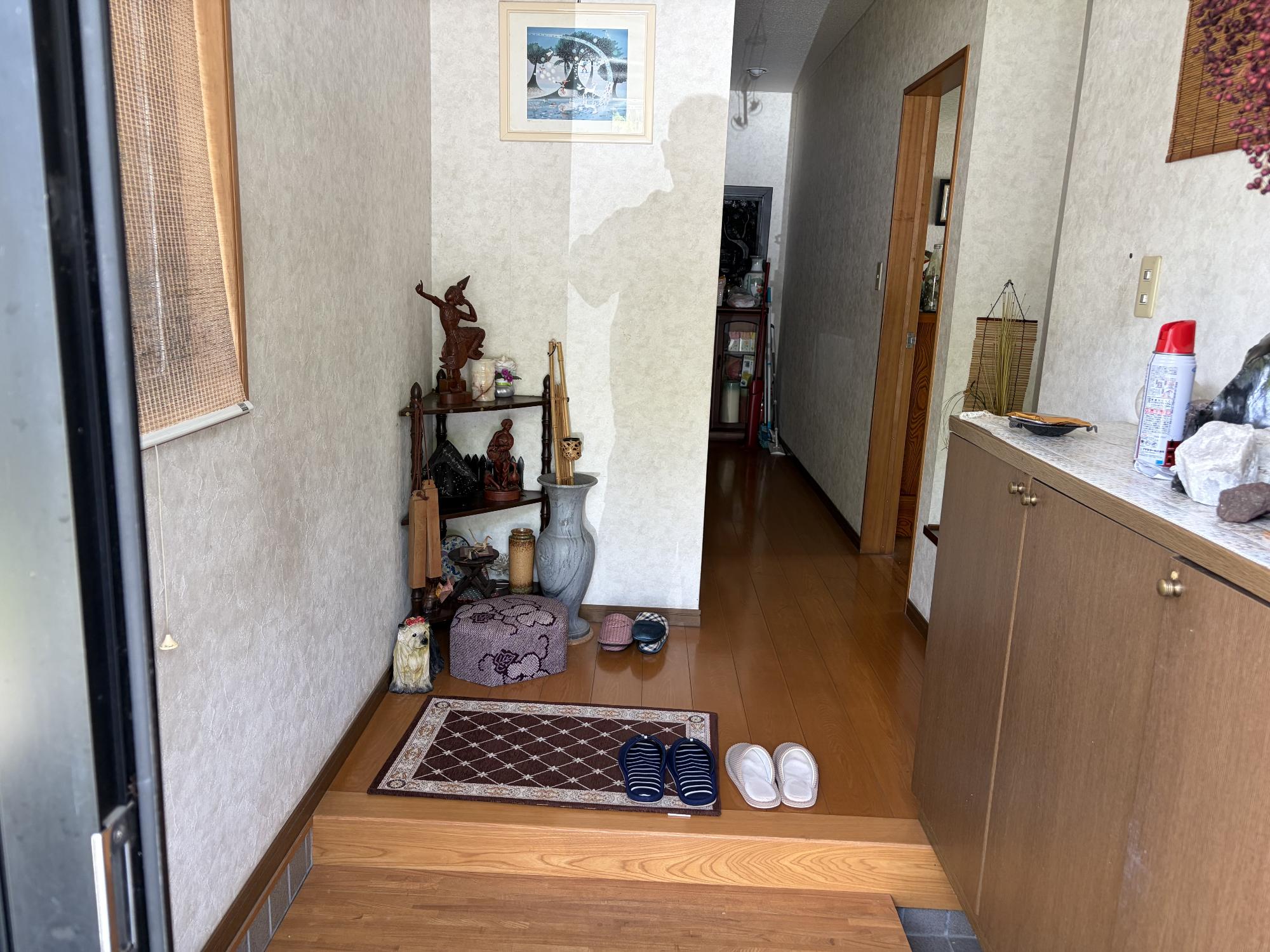 左側に出窓、右側に下駄箱、中央正面上部に壁かけ、正面に花瓶、飾り棚、手前に玄関マット、スリッパがある玄関内観の写真