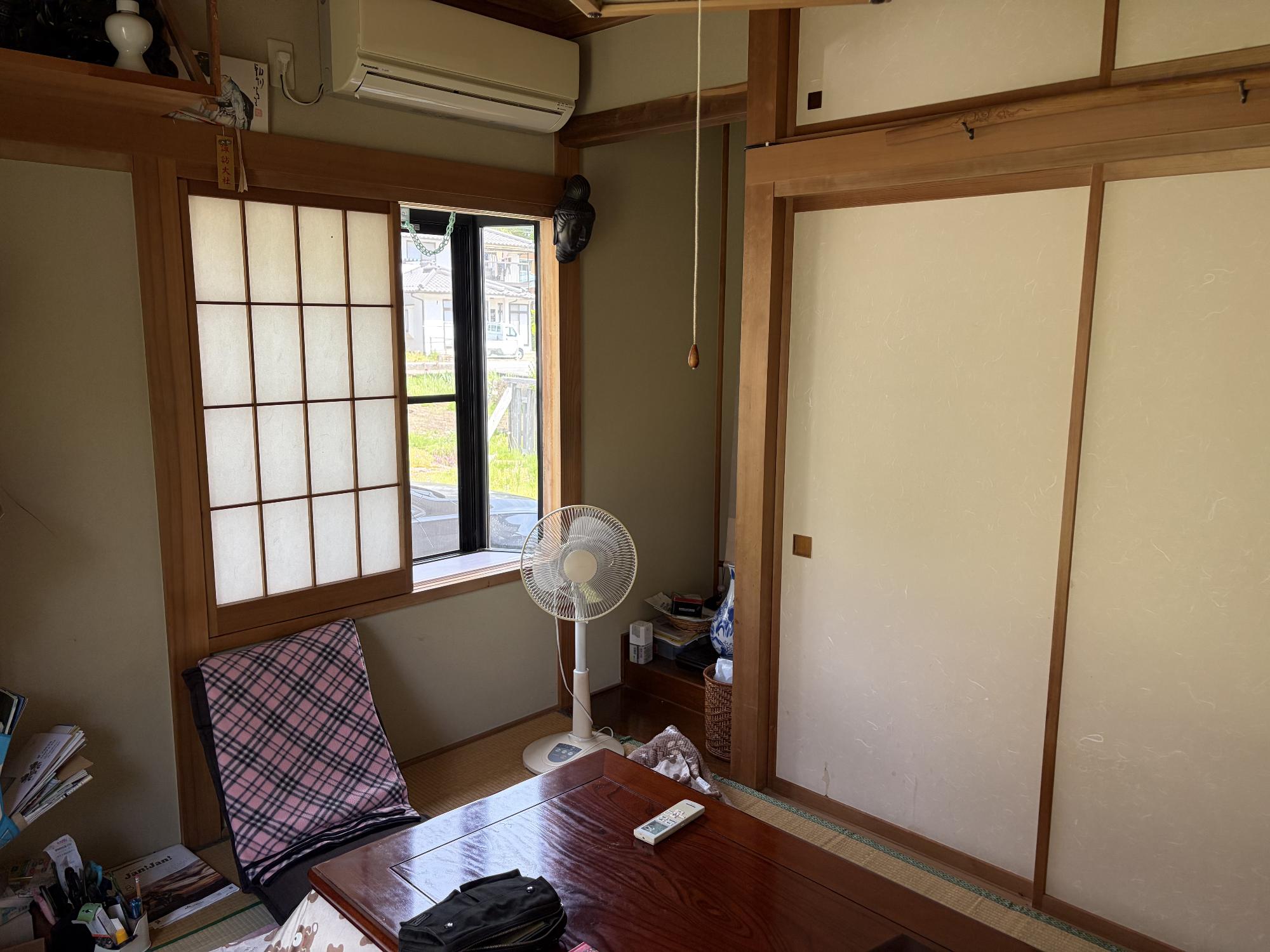 正面奥に床の間、その右側に障子戸が開いた出窓、正面上部にエアコン、正面下部に扇風機、座卓、右側に押し入れ引戸がある6帖和室の内観写真
