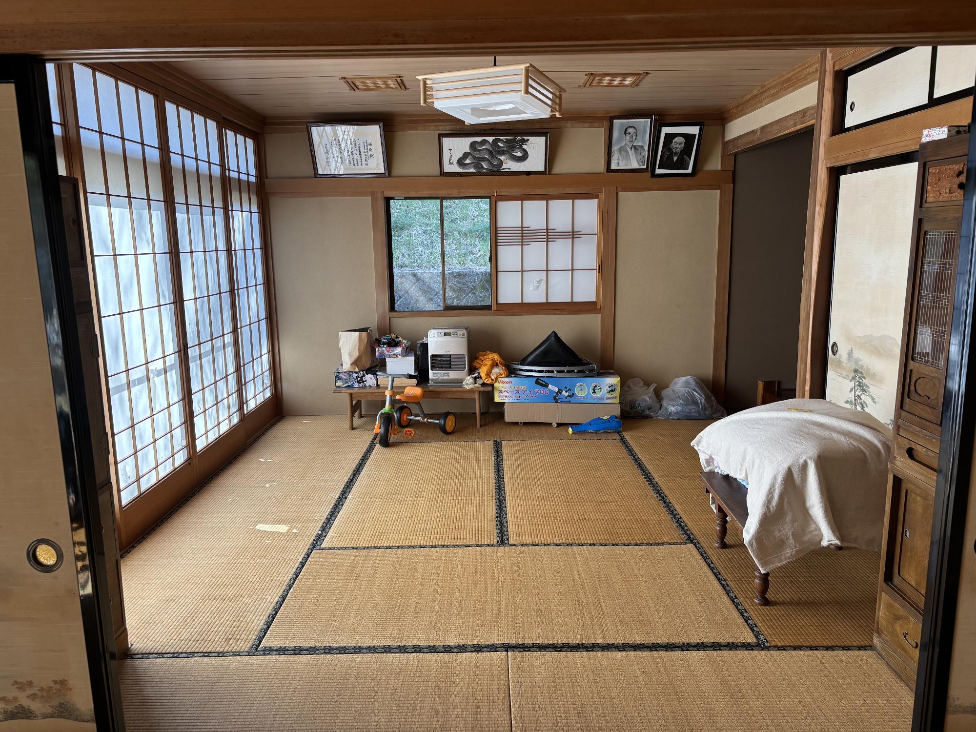 左側に障子引戸、右側に床の間、押入れ、正面中央に障子引戸を開いたアルミサッシ戸、その手前に玩具、上部に額に入った飾り物、正面中央上部に照明器具がある、10帖和室の内観写真