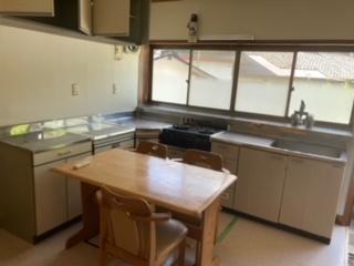 左側に調理台、中央奥にガスコンロ、右側にシンク、中央奥上部にサッシ、中央手前に食卓机、椅子がある台所内観写真