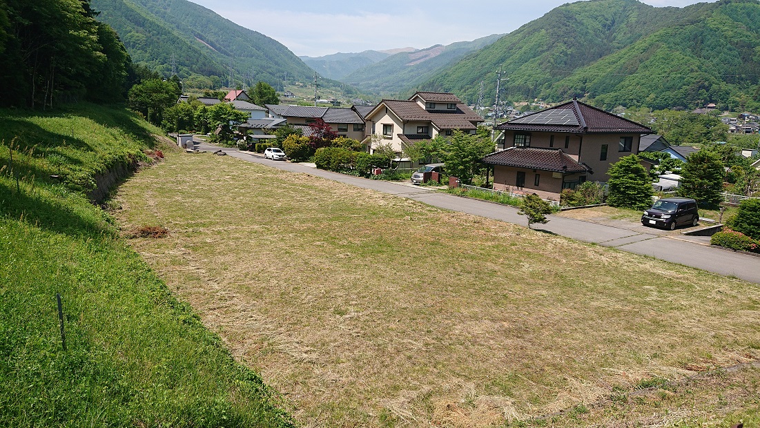 奥に山々が見える住宅地の道路の向かい側にある分譲地を高い場所から撮影した写真